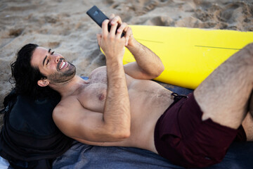 Handsome man resting on the beach, using the phone. Surfer taking a break on the beach..