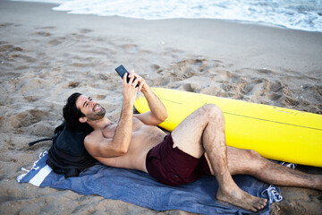 Handsome man resting on the beach, using the phone. Surfer taking a break on the beach..
