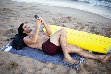 Handsome man resting on the beach, using the phone. Surfer taking a break on the beach..