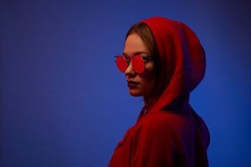 Young model posing in a studio in a trendy neon light. Woman in round sunglasses and hoodie with colorful light effects.