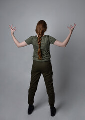 Full length portrait of pretty red haired woman wearing army green khaki shirt, utilitarian pants and boots. Standing pose with back to the camera, isolated on studio background.