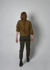 Full length portrait of pretty red haired woman wearing army green khaki shirt, utilitarian pants and boots. Standing pose with back to the camera, isolated on studio background.