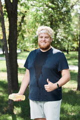 Naklejka na ściany i meble Portrait of happy overweight man with bottle of water smiling at camera and showing thumb up after training in the park