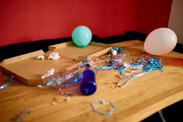Messy wooden table with pizza box, empty blue bottles, wine glass and party decorations at morning...