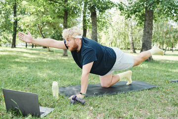 Overweight man exercising with dumbbells on exercise mat in the park and watching sports training online on laptop
