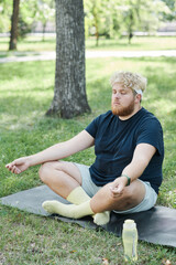 Naklejka na ściany i meble Overweight man sitting on the grass with eyes closed and concentrating on yoga exercises in the park