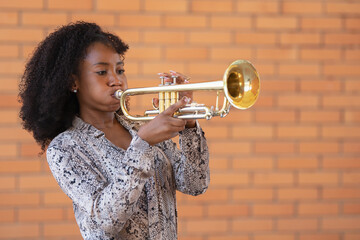 Portrait of a young afro american woman playing the trumpet outside on a brick wall background - obrazy, fototapety, plakaty