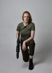 Full length portrait of pretty red haired woman wearing army green khaki shirt, utilitarian pants and boots. Holding a gun as weapon, isolated on studio background.