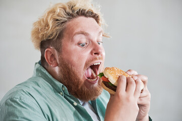 Close-up of overweight man opening his mouth and eating big burger with pleasure isolated on white...