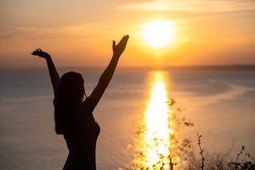 Fototapeta premium silhouette of a person with arms raised, successful young woman standing in the sunset celebrating success