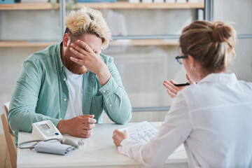 Upset overweight man sitting at the table and covering his face with hand he listening to diagnosis of the doctor at hospital