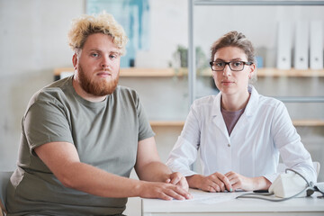 Fototapeta premium Portrait of young overweight man sitting at the table with his nutritionist and they looking at camera together at office
