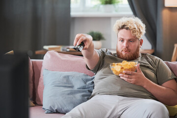 Lazy overweight man sitting on sofa with chips and using remote control to watch TV at home