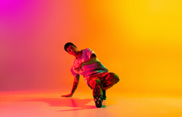 Fototapeta na wymiar Full length portrait of young stylish man, break dancing dancer in casual clothes isolated over gradient pink yellow background at dance hall in neon light.