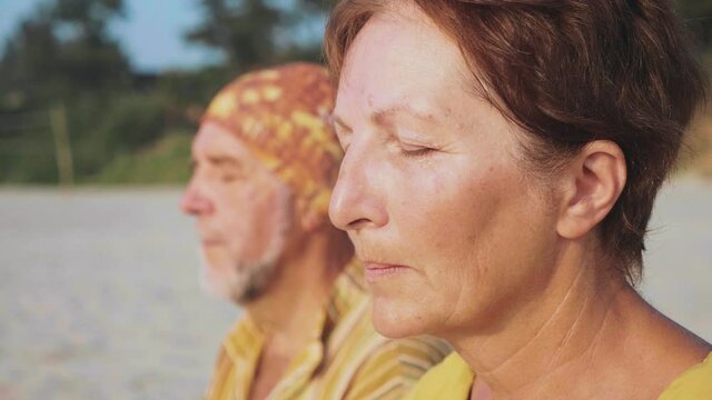 Senior couple sits and meditating together on sandy beach. Elderly man and woman sits in turkish pose with closed eyes, female portrait.