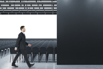 Businessman standing in modern grey auditorium with seating, empty screen and mock up place for...