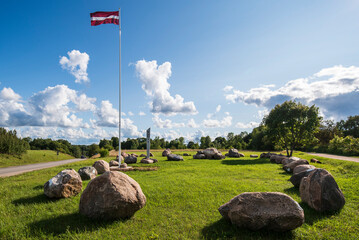 Memorial place with large stones. A flowing Latvian flag on a long mast. - obrazy, fototapety, plakaty
