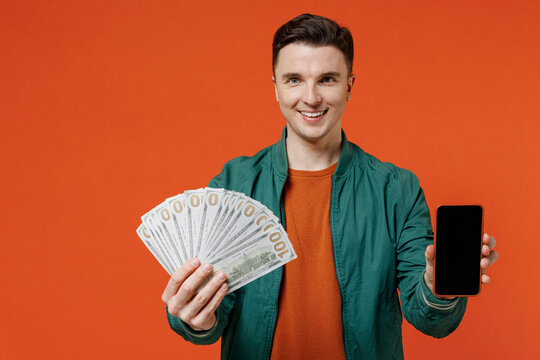 Young brunet man 20s wear red t-shirt green jacket hold in hand use mobile cell phone with blank screen workspace area fan of cash money in dollars isolated on plain orange background studio portrait