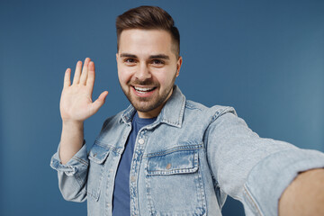 Close up happy fun young brunet man 20s wears denim jacket doing selfie shot on mobile phone meet greet waving hand isolated on dark blue background studio portrait. People emotions lifestyle concept.