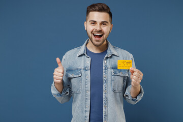 Satisfied happy excited young brunet man 20s wears denim jacket hold in hand credit bank card show...