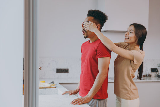 Young couple two woman american african man in casual clothes wife close husband eyes with arms play guess who or hide and seek in kitchen at home together Healthy diet lifestyle relationship concept