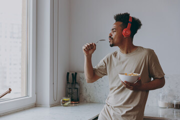 Young african american man 20s in casual clothes headphones listen music eat breakfast muesli cereals with milk fruit in bowl sing song in spoon microphone prepare cook food in kitchen at home indoor.