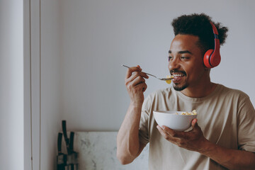 Young smiling african american man 20s in casual clothes headphones listen music eat breakfast muesli cereals with milk fruit in bowl prepare cook food in kitchen at home indoor. Healthy diet concept