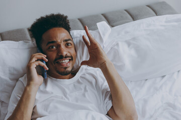 Young irritated awaken african american man in nightwear talk mobile cell phone lying in bed rest relax spend time in bedroom lounge home in own room house wake up dream be lost in reverie good day