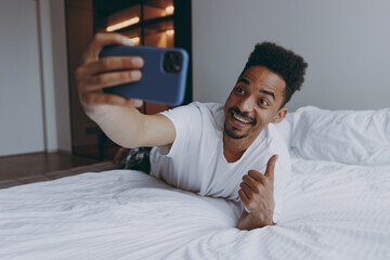 Full length fun young african american man in nightwear do selfie shot on mobile cell phone show thumb up gesture lying in bed rest relax spend time in bedroom lounge home in own room house wake up.