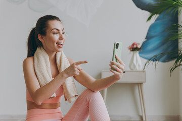 Young strong sporty athletic fitness trainer instructor woman in pink tracksuit towel doing yoga exercises do selfie shot point index finger on mobile cell phone sit on mat floor at home gym indoor.