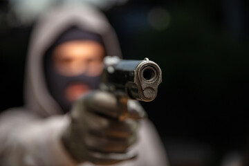 Man aiming with a pistol, dark outdoors background, closeup view. Hooded thief holding a pistol, street at night background,