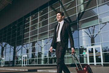 Bottom view satisfied fun young traveler businessman man wear black dinner suit walk go outside at international airport terminal with suitcase valise crossing road Air flight business trip concept