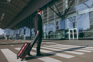 Side profile view full body young traveler businessman man in black suit facial mask walk outside at international airport terminal with suitcase valise crossing road Air flight business trip concept.