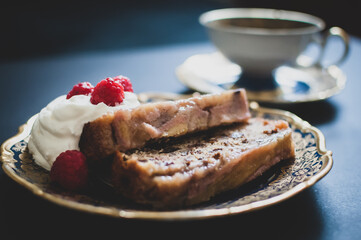 Delicious sweet baked homemade banana bread with nuts, raisins, whipped cream and raspberries on small dessert ornamental plate. A cup of tea in the background.  Vintage design. Selective focus.