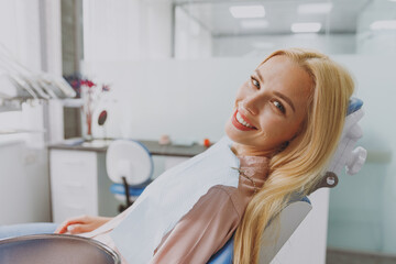 Close up young smiling happy blonde woman 20s covered by napkin sitting at dental office chair indoor light modern cabinet waiting stomatologist for oral procedure. Healthcare caries enamel treatment