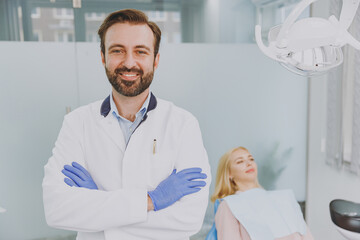 Obraz premium Young confident smiling fun male man doctor hold hands crossed folded stand near patient woman sitting at dentist office chair indoor light cabinet near stomatologist Healthcare oral enamel treatment.