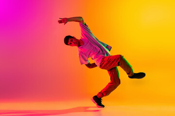 Fototapeta na wymiar Portrait of young stylish man, break dancing dancer training in casual clothes isolated over gradient pink yellow background at dance hall in neon light.