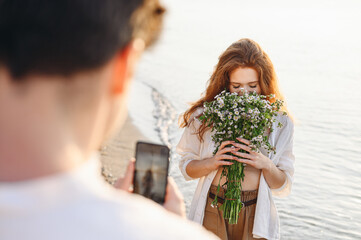 Happy young couple family man woman in white clothes sniff flowers bouquet take photo on mobile cell phone rest together at sunrise over sea beach ocean outdoor seaside in summer day sunset evening.
