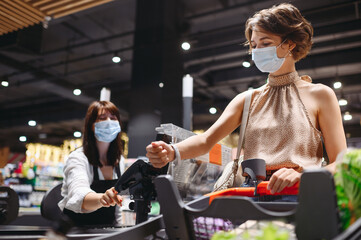 Obraz premium Young caucasian woman 20s in casual clothes mask shopping at supermaket with grocery cart put smart watch to modern bank payment terminal process acquire payments near cashier checkout inside store.