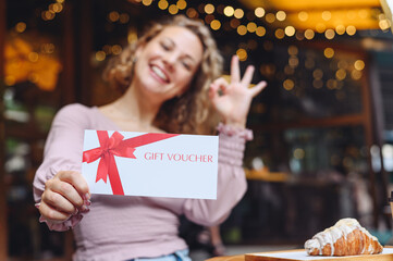 Young happy woman in casual clothes sit at cafe table eat breakfast show ok gesture hold gift certificate coupon voucher card for store relaxing in restaurant during free time indoors. Focus on card. - obrazy, fototapety, plakaty