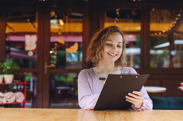 Fototapeta premium Young happy smiling waiter businesswoman woman wear casual clothes look aside standing at coffee shop cafe hold black clipboard with papers reading document write menu working in restaurant indoors