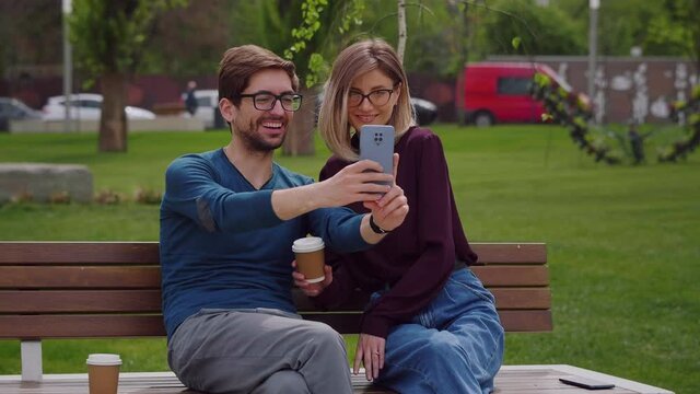 Two friends with glasses, pretty woman and handsome man sit on the bench outside take selfie with phone, feel happy smiling. Social networking outdoors.