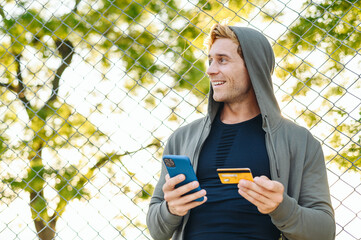 Young happy fun wistful man 20s wear grey sportswear hood look aside using mobile cell phone hold credit bank card doing online shopping order delivery booking tour Outdoor courtyard sports concept