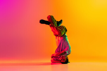 In action. Young man, break dancing dancer ptacticing in casual clothes isolated over gradient pink yellow background at dance hall in neon light.