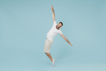 Full length young smiling happy man 20s in white t-shirt leaning back with outstretched hand dancing fooling around stand on toes isolated on plain pastel light blue color background studio portrait