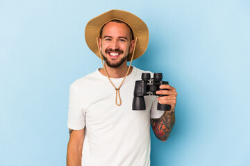 Young caucasian man with tattoos holding binoculars isolated on blue background  happy, smiling and cheerful.