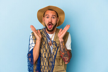 Young caucasian fisherman with tattoos holding net isolated on blue background  surprised and shocked.
