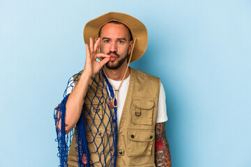 Young caucasian fisherman with tattoos holding net isolated on blue background  with fingers on lips keeping a secret.