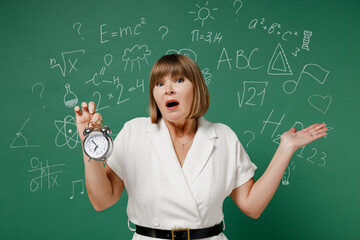 Amazed puzzled teacher mature elderly lady woman 55 wear white shirt hold clock spread hands isolated on green wall chalk blackboard background studio. Education in high school September 1 concept.