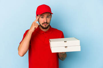 Young caucasian delivery man with tattoos holding pizzas isolated on blue background  pointing temple with finger, thinking, focused on a task.